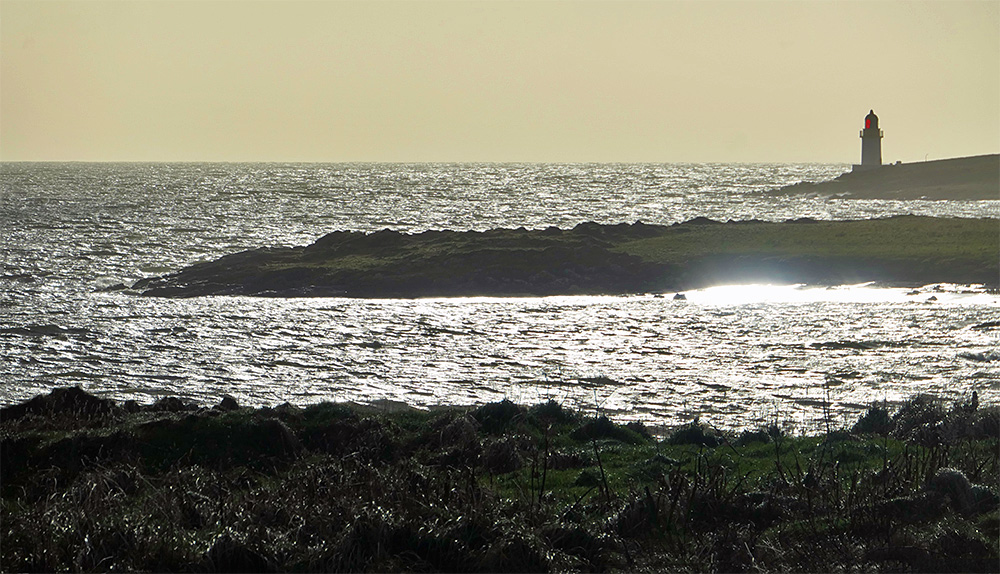 Picture of a sea loch with sun reflections along the shore, a small lighthouse in the distance