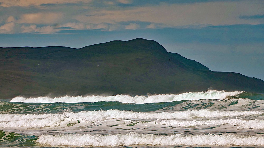Picture of waves rolling into a wide bay, hills above cliffs in the background