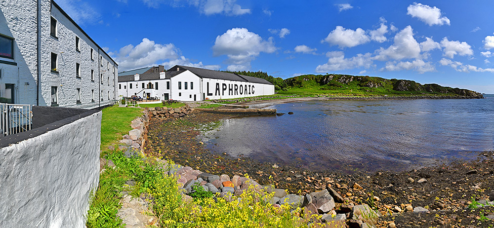Panoramic picture of Laphroaig distillery on Islay on a bright June afternoon, some yellow flowers below, blue skies with fluffy white clouds above