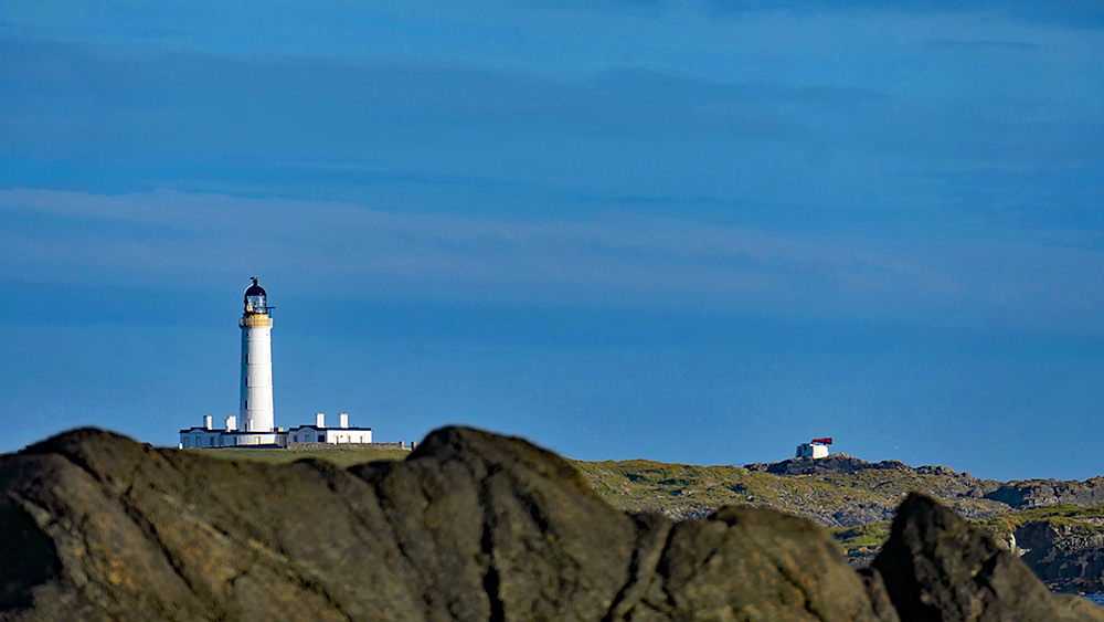 Picture of a lighthouse seen from behind some distant coastal rocks
